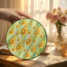 Load image into Gallery viewer, Hand displaying the Make It Rain stained glass-style suncatcher with golden raindrops and fresh green artwork in a softly lit room with flowers.