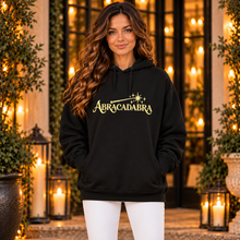 Ladda upp bild till gallerivisning, Woman wearing black ABRACADABRA organic cotton hoodie with yellow wand graphic and white jeans, standing outside a warmly lit entrance.