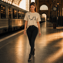 Load image into Gallery viewer, A woman walking on a train station platform wearing a beige graphic t-shirt with the word 'DISCRED' printed in black, paired with black pants and black boots.