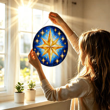 Ladda upp bild till gallerivisning, Woman holding the Written in the Stars glass suncatcher toward a sunlit window, showcasing its radiant golden stars and deep blue artwork.