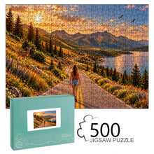Load image into Gallery viewer, The Road Ahead 500 piece wooden jigsaw puzzle featuring a woman walking down a scenic mountain road with wildflowers, deer, soaring birds, a lake, pine trees, mountains, and a golden sunset.