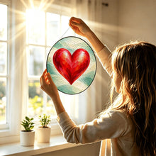 Carica l'immagine nel visualizzatore di Gallery, Woman holding the Follow Your Heart red heart stained glass-style suncatcher toward a bright window in natural sunlight.
