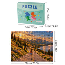 Load image into Gallery viewer, The Road Ahead 500 piece wooden jigsaw puzzle featuring a woman walking down a scenic mountain road with wildflowers, deer, soaring birds, a lake, pine trees, mountains, and a golden sunset.
