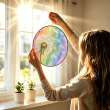Carica l'immagine nel visualizzatore di Gallery, Woman holding the Wish Come True dandelion glass suncatcher toward a sunlit window, showcasing its colorful pastel rainbow design.
