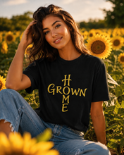 Load image into Gallery viewer, A woman sitting in a sunflower field wearing a black graphic t-shirt with 'GROWN ME' printed in yellow, paired with blue jeans.
