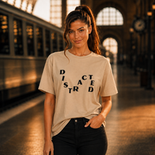 Load image into Gallery viewer, A woman stands on a train station platform wearing a beige crewneck t-shirt with the word 'DISRUPTED' printed in black across the front, paired with black jeans.
