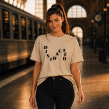 Load image into Gallery viewer, A woman stands on a train platform wearing a beige graphic t-shirt with the word 'DISRACTED' spelled out in black letters, paired with black pants.
