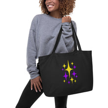 Carica l'immagine nel visualizzatore di Gallery, Model carrying Astro Glow oversized black organic cotton tote bag featuring original purple and yellow cosmic starburst artwork from Little Yellow Bungalow.
